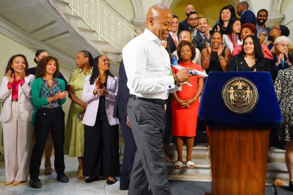 Mayor Eric Adams standing at a podium with crowd, holding a small airplane, celebrating a handshake agreement on the 2025 NYC budget with City Council Speaker Adrienne Adams