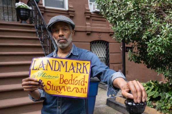 A black man in a blue shirt and cap holds a sign that says, "Landmark Historic Bedford-Stuyvesant," as he stands in front of brownstone steps.