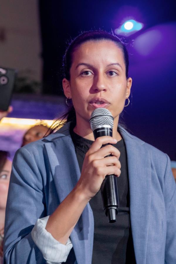 Queens City Councilwoman Tiffany Caban, wearing a black shirt and blue suit jacket, speaking into a microphone.