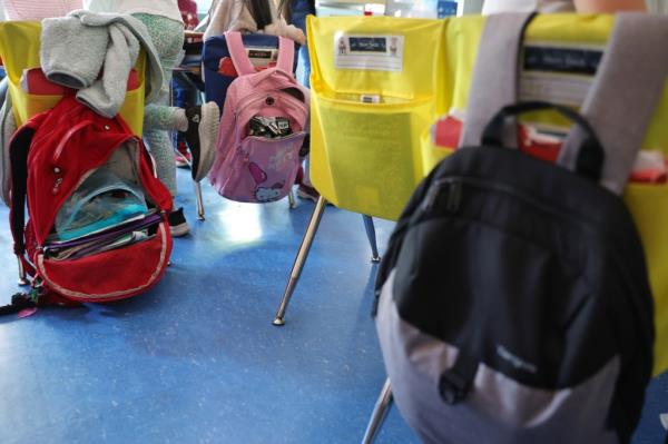 A stock photo of kids' backpacks in a classroom hanging on the backs of chairs